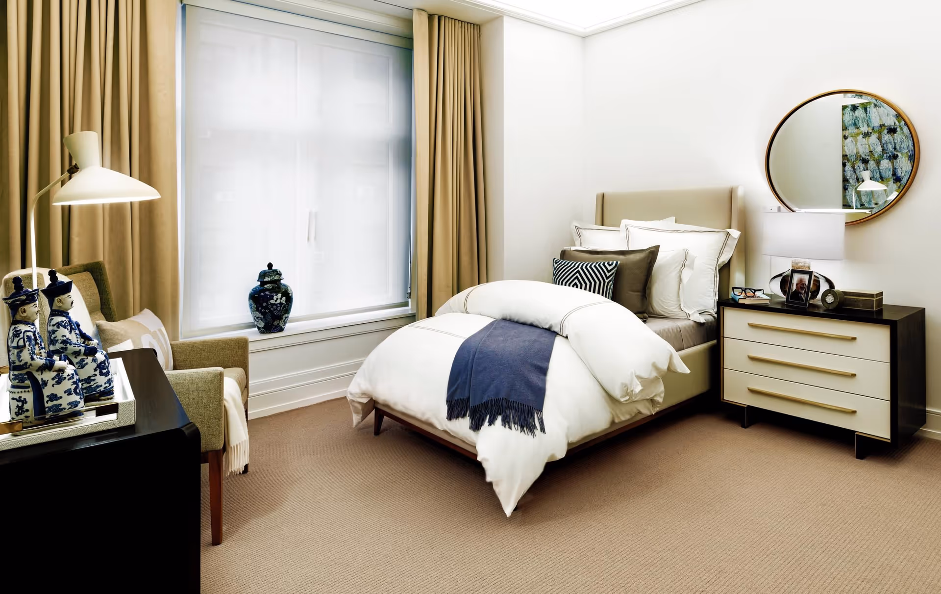 A neatly made bed with white and brown pillows and a blue throw blanket in a bright bedroom. There is a beige armchair with a pillow and a white floor lamp next to a window with beige curtains. A dark wooden dresser with three drawers and a round mirror above it is on the right side of the bed. Decorative blue and white ceramic figurines and a vase are placed on furniture near the window.