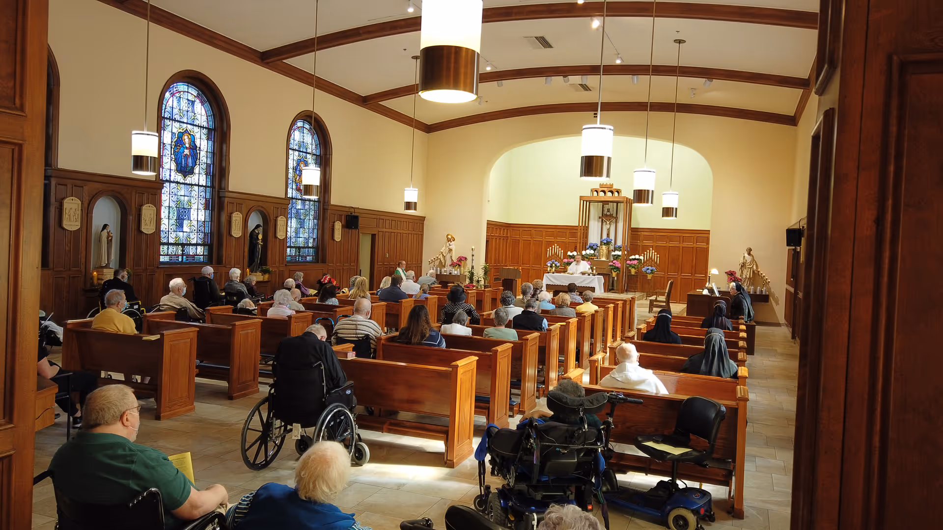 Interior view of a chapel with wooden pews filled with elderly people, some in wheelchairs, attending a service. The chapel features stained glass windows, religious statues, and hanging pendant lights.