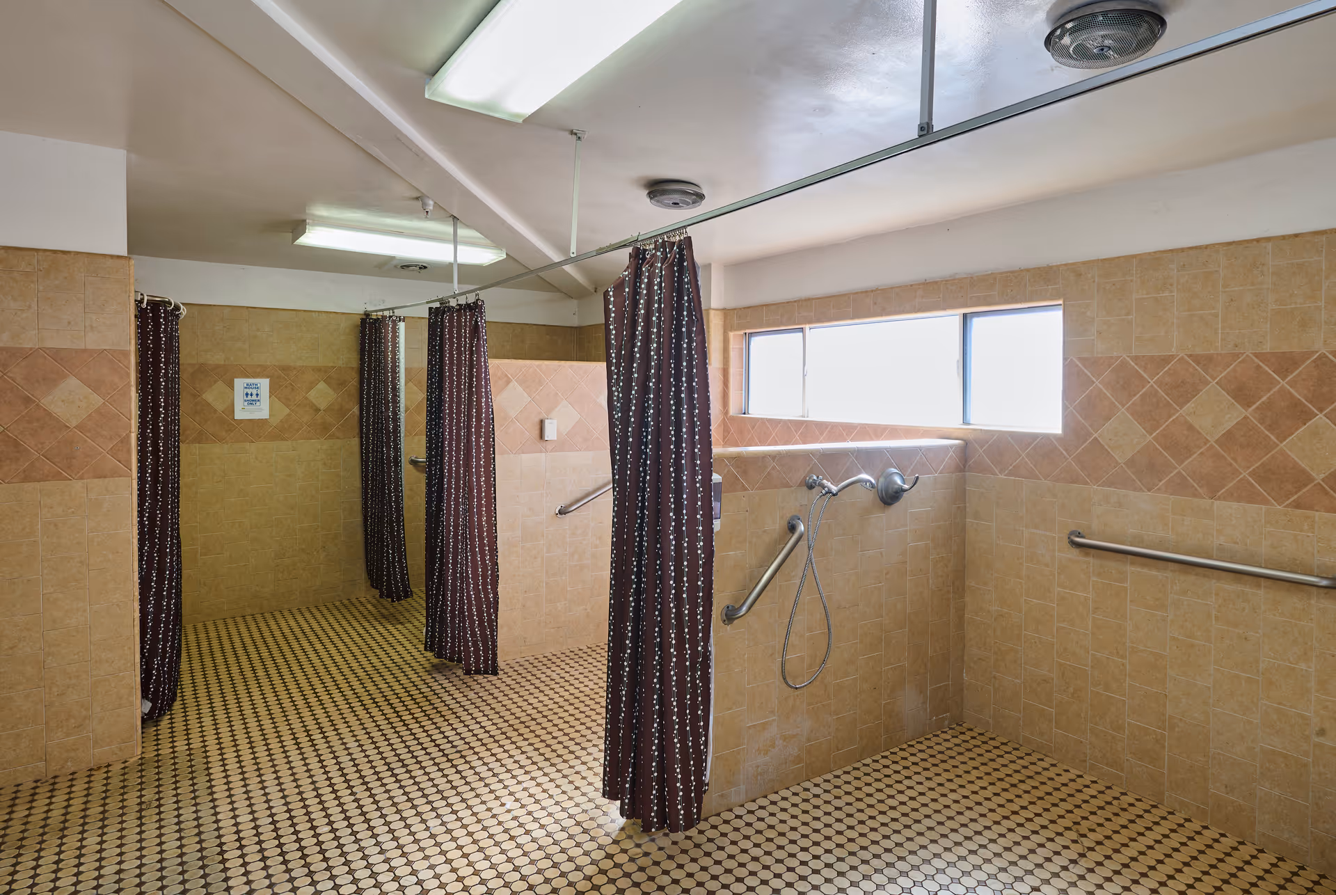 A communal shower area with multiple shower stalls separated by brown patterned curtains. The walls are tiled with beige and brown tiles, and the floor has small round tiles in a beige and brown pattern. There are grab bars installed on the walls for support, and a window lets in natural light.