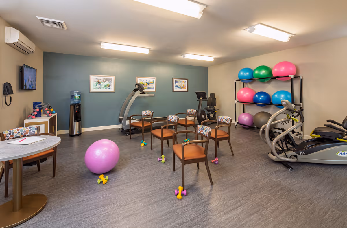 A fitness room with exercise equipment including a treadmill, stationary bike, and elliptical machine. Several colorful exercise balls are stored on a rack against the wall. There are six chairs arranged in a circle with small dumbbells placed near them. A pink exercise ball and more dumbbells are on the floor. The room has a water cooler, a small table with a chair, and framed artwork on the walls.