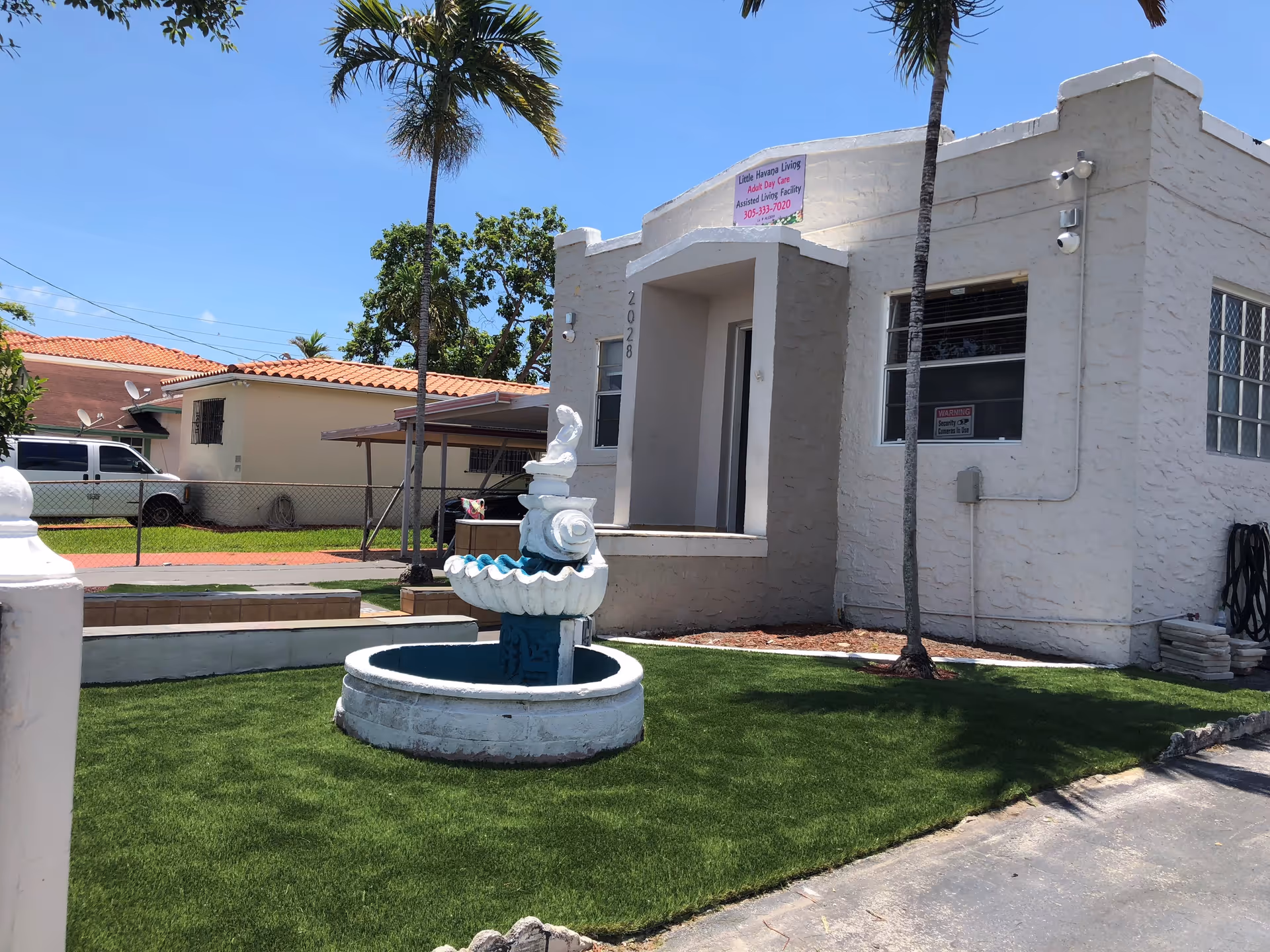 Exterior view of a single-story building with a textured white facade and a small covered entrance. In front of the building is a green lawn with a decorative white and blue fountain. Two tall palm trees are visible near the building. A neighboring house and a white van are seen in the background under a clear blue sky.