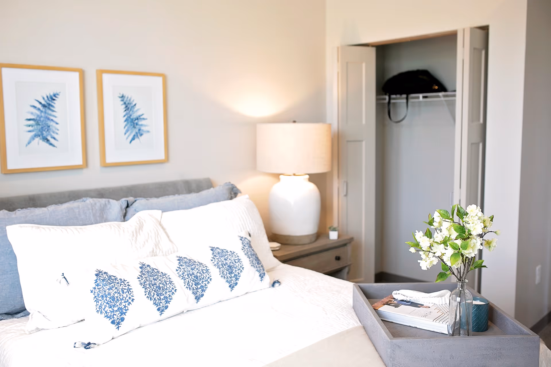 A bright and cozy bedroom featuring a neatly made bed with white and blue patterned pillows, a wooden nightstand with a white lamp, two framed botanical prints on the wall, an open closet with a bag on the shelf, and a tray with a vase of white flowers and a book on the bed.