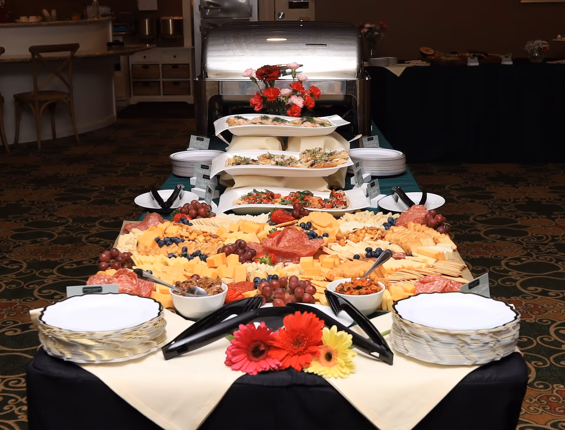 A buffet table set up with a variety of cheeses, crackers, grapes, and sliced meats arranged on platters. There are also small bowls with spreads or dips, stacks of white plates with gold rims, and serving tongs. The table is decorated with colorful flowers and there is a chafing dish with additional food in the background in a dining area.