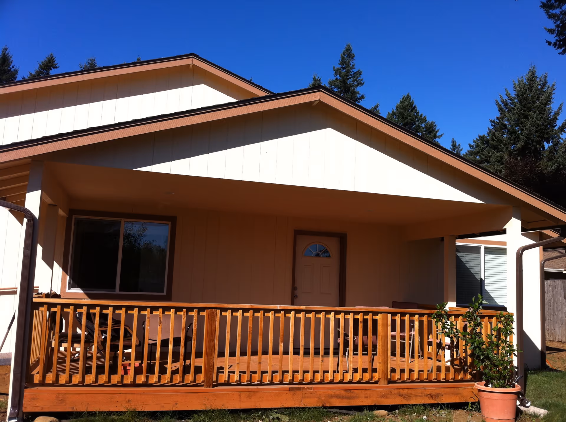 Front view of a single-story house with a covered wooden porch. The porch has a wooden railing and several chairs. There is a door and two windows visible on the house exterior. A potted plant is placed near the porch. Tall evergreen trees are in the background under a clear blue sky.
