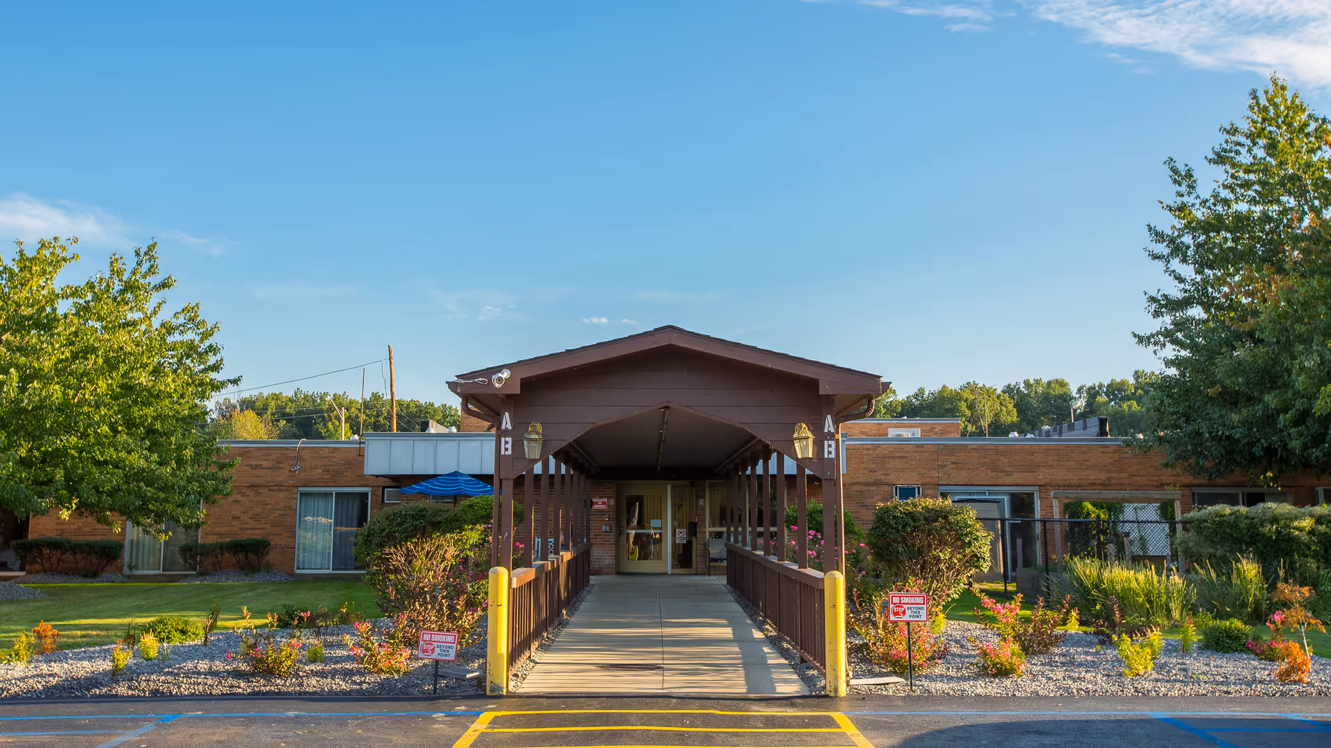 Front exterior view of Carington Park facility showing a covered walkway leading to the entrance, surrounded by landscaped gardens with bushes and trees under a clear blue sky.