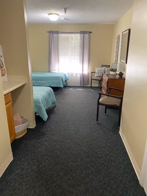 A memory care facility room with two twin beds covered in light blue bedspreads, a window with sheer curtains letting in natural light, a wooden dresser with decorative items, and two chairs placed near the dresser. The room has beige walls and dark carpet flooring.