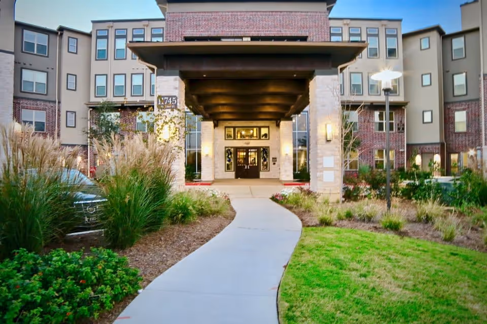 Front exterior view of a multi-story senior living facility with a covered entrance supported by stone pillars, landscaped greenery, and a curved sidewalk leading to the entrance.