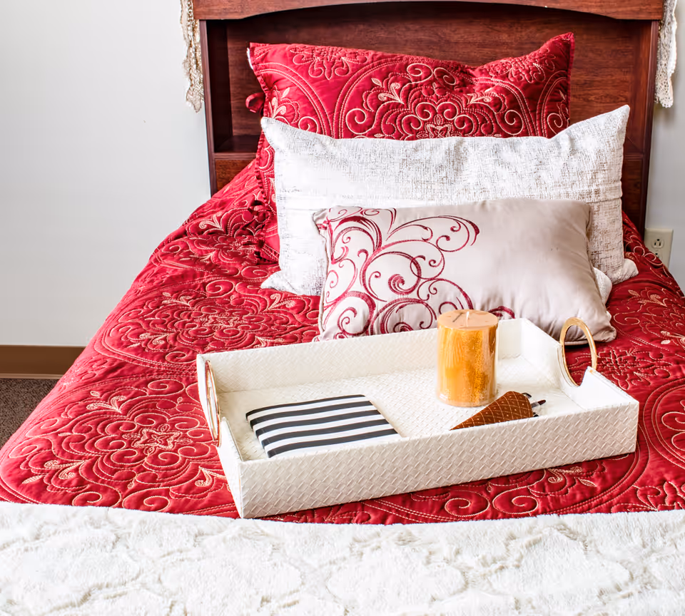 A neatly made bed with a red quilted bedspread featuring intricate patterns, three decorative pillows in red and beige tones, and a white textured tray on top holding a striped notebook, a brown cone-shaped object, and a large orange candle.