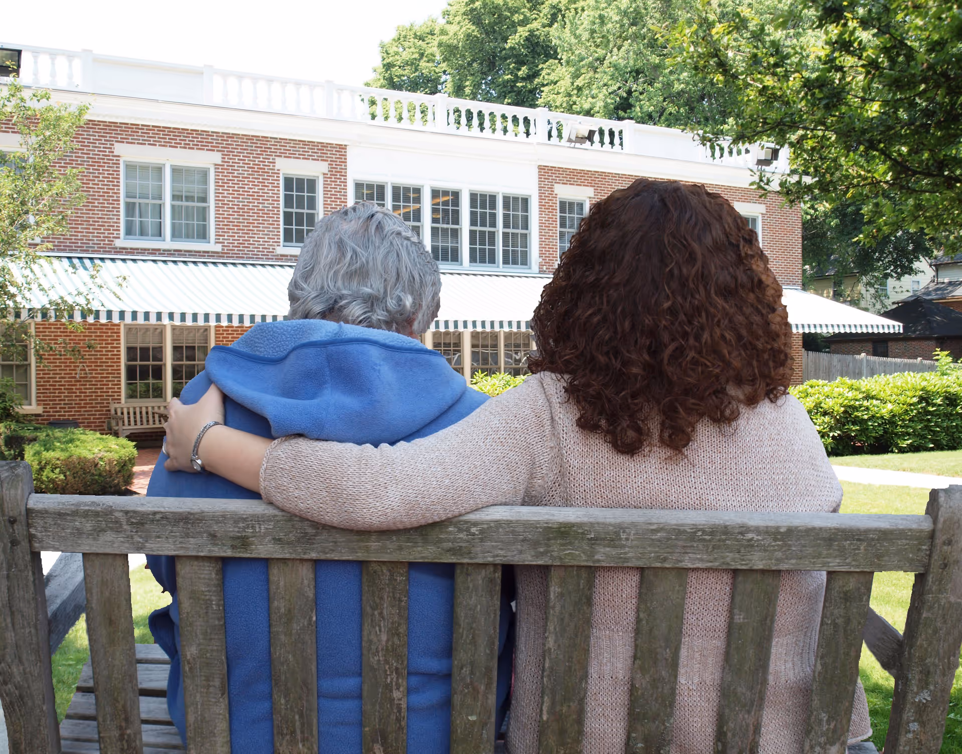 An elderly person with gray hair wearing a blue jacket sits on a wooden bench outdoors with a younger person with curly brown hair and a beige sweater. The younger person has their arm around the elderly person. They are facing a large brick building with white trim and multiple windows, surrounded by greenery and trees.