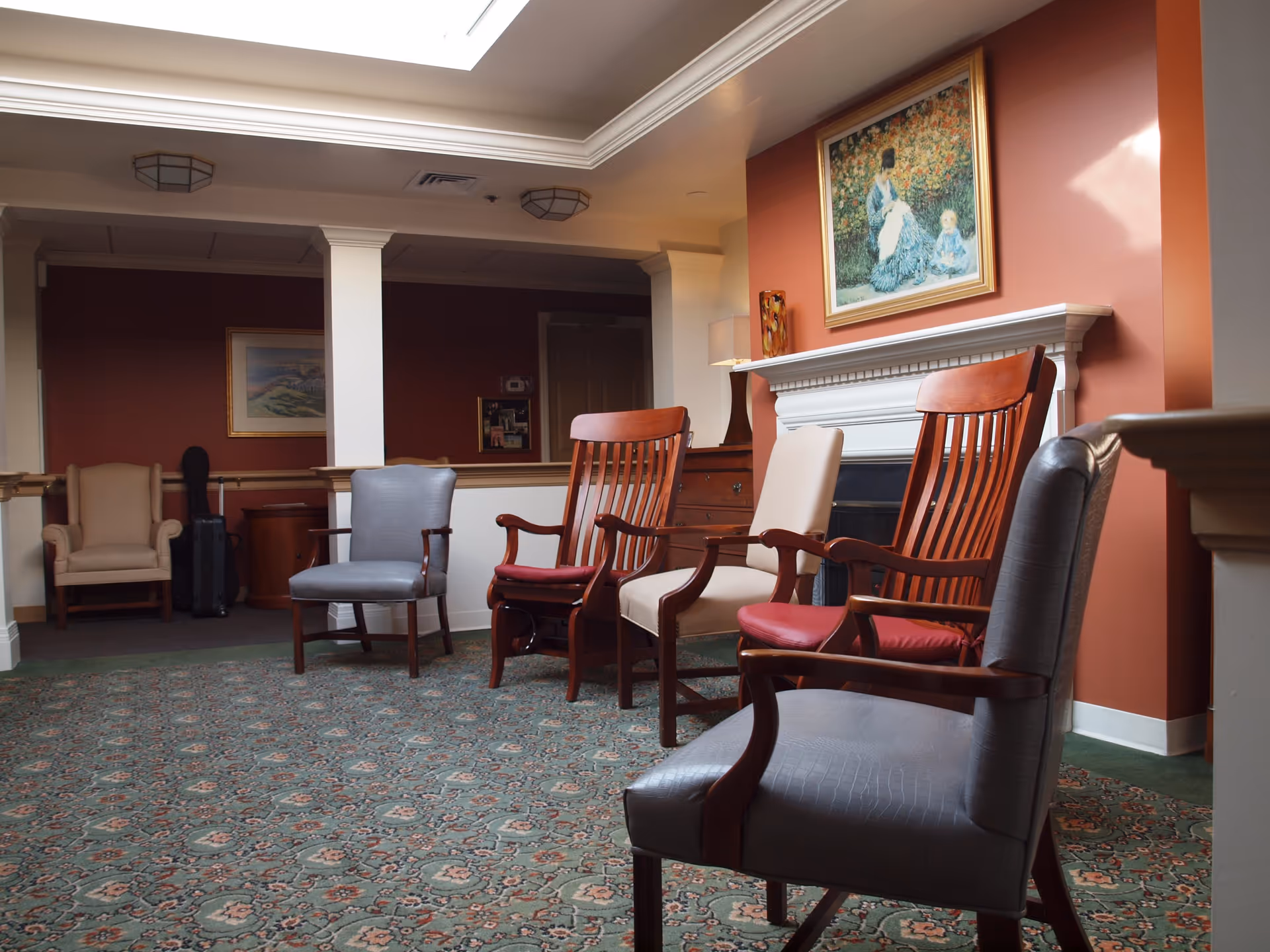 A cozy sitting area in a memory care assisted living facility featuring a variety of wooden and upholstered chairs arranged on a patterned carpet. The walls are painted a warm reddish color, and there is a white fireplace with a framed painting above it. The room is softly lit with natural light from a skylight and a table lamp on a wooden side table.