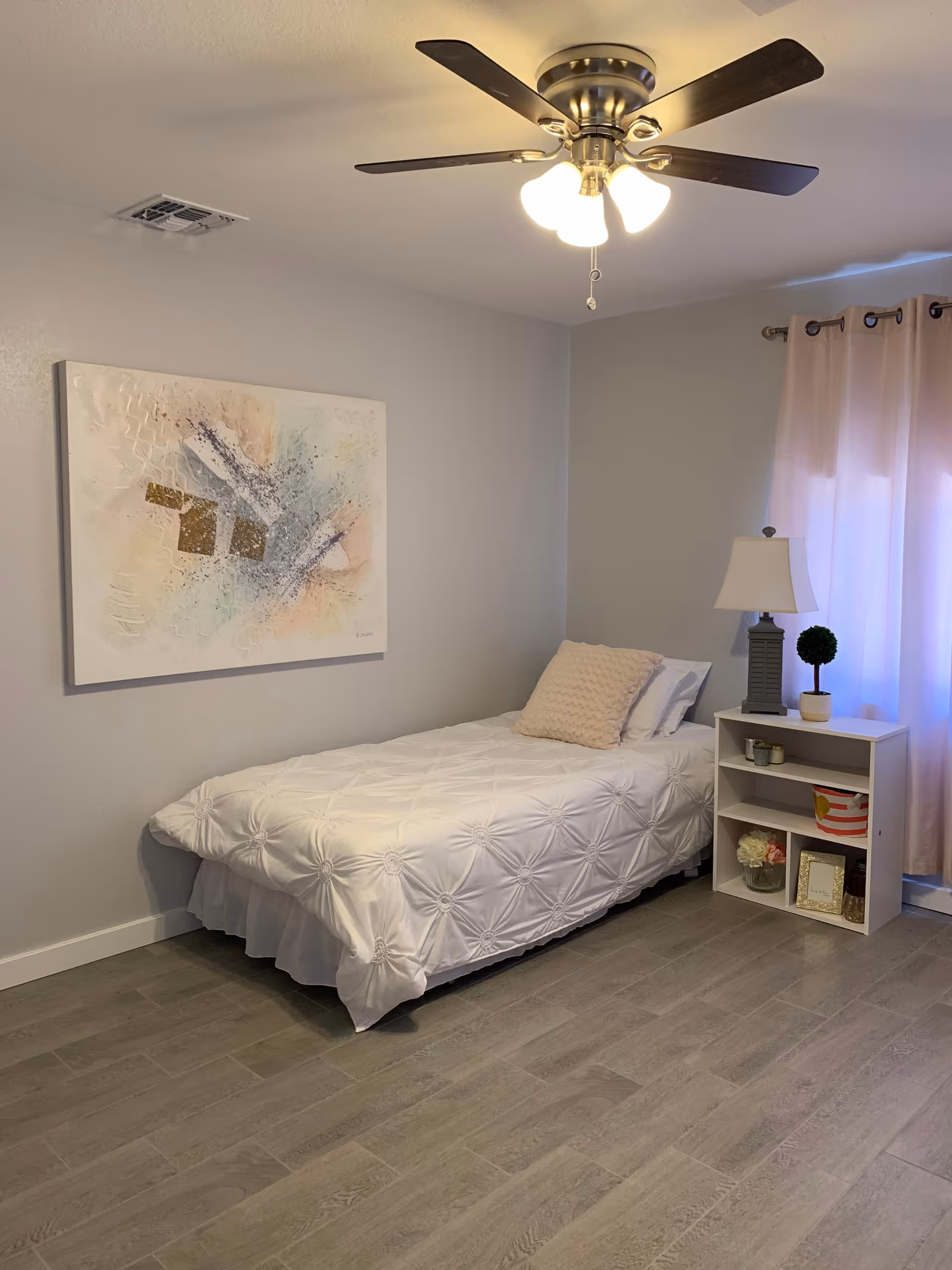 A simple bedroom with a single bed covered in a white textured comforter and a beige pillow. Next to the bed is a small white nightstand with a lamp, a small potted plant, and decorative items. A large abstract painting hangs on the wall above the bed. The room has light-colored walls, wood-look flooring, a ceiling fan with lights, and a window with light pink curtains.
