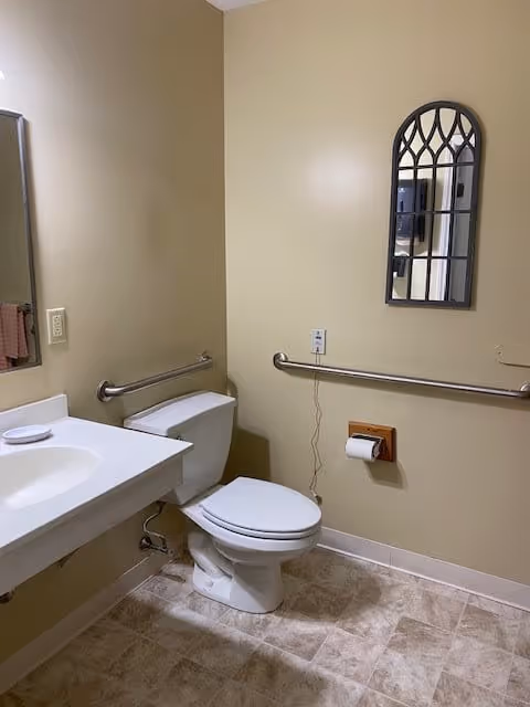 A clean bathroom with beige walls and tiled floor. It features a white toilet with grab bars on the wall behind and beside it. To the left is a white sink with a mirror above it. A decorative arched window-style mirror hangs on the wall above the toilet paper holder.
