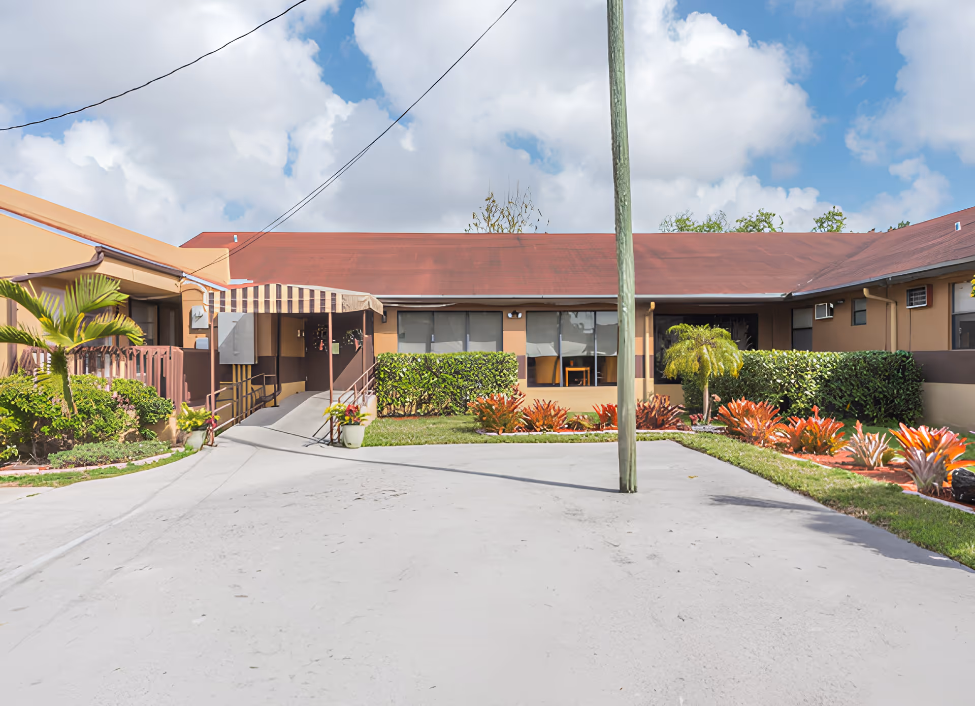 Exterior view of a single-story building with a red roof and beige walls, featuring a wheelchair accessible ramp with a striped awning. The building is surrounded by neatly trimmed bushes, colorful plants, and a paved driveway under a partly cloudy sky.