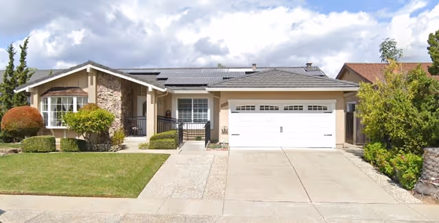 Front exterior view of a single-story house with a two-car garage, stone and beige siding, solar panels on the roof, a well-maintained lawn, and some bushes and trees under a partly cloudy sky.