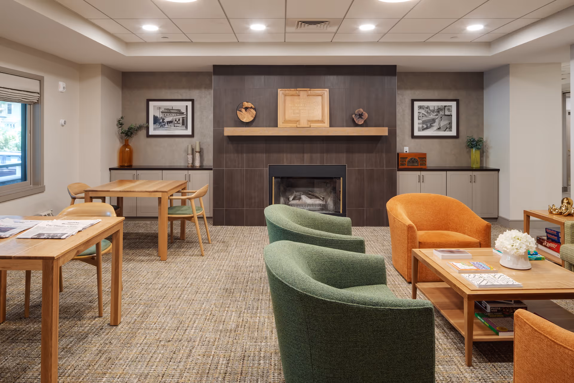A cozy common area in a senior living facility featuring a fireplace with a wooden mantel, two green armchairs, two orange armchairs, and wooden tables with chairs. The room has a carpeted floor, framed black and white photos on the walls, and decorative items including vases and books on the tables and cabinets.