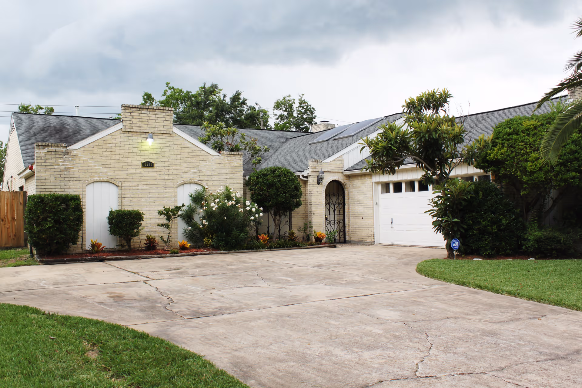 Exterior view of a single-story brick house with a large driveway, a garage door, and various shrubs and trees in the front yard under a cloudy sky.