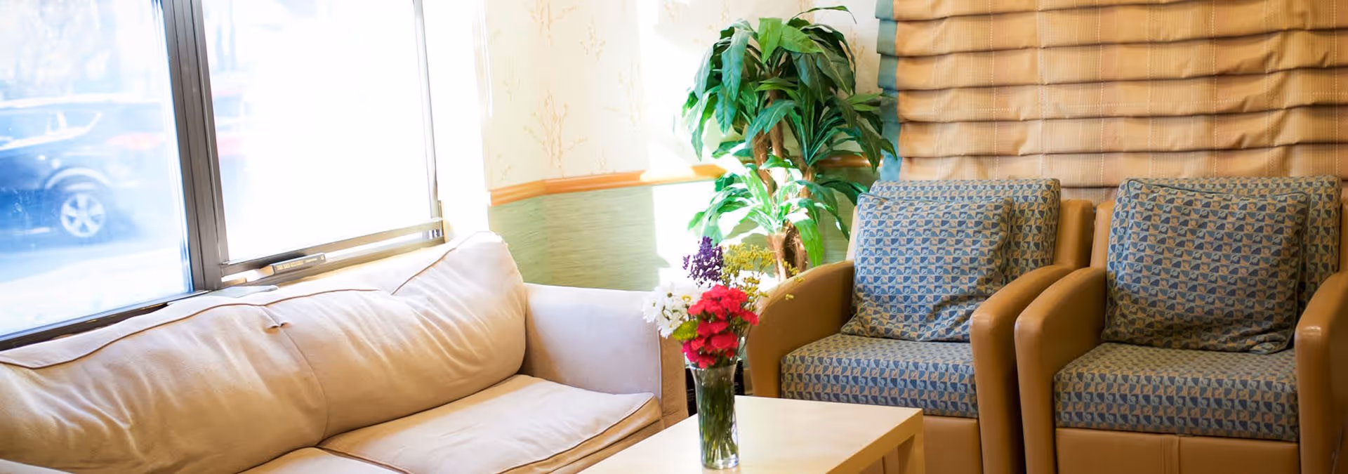 A bright and cozy seating area with a beige couch on the left and two patterned armchairs on the right. A small wooden table in front holds a vase with colorful flowers. There is a large window letting in natural light and a green potted plant in the corner.