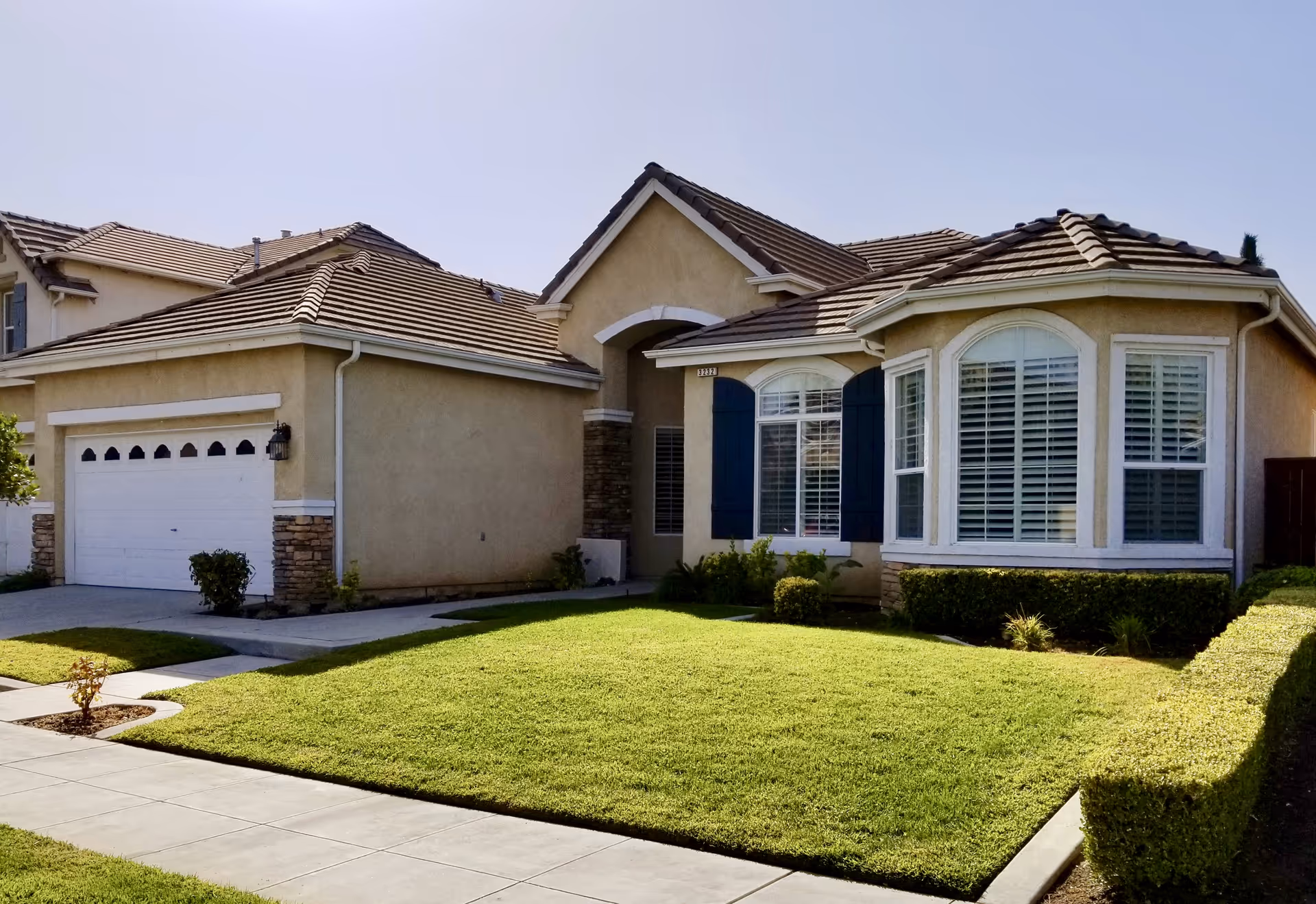 Exterior front view of a single-story residential-style building with a well-maintained green lawn, a concrete driveway leading to a two-car garage, and windows with white shutters and blue trim.