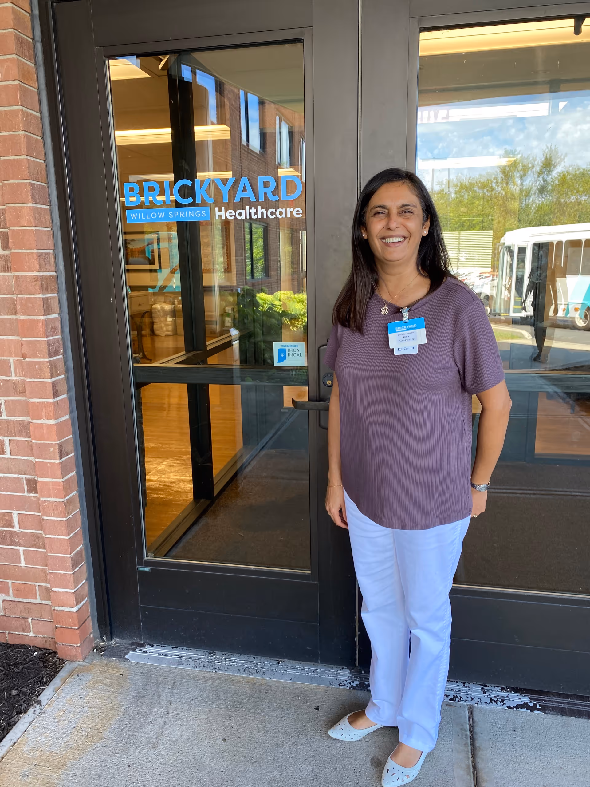 A woman wearing a purple top and white pants stands smiling in front of glass doors with the sign 'Brickyard Willow Springs Healthcare'. The entrance shows a brick wall on the left and a reflection of a bus and trees on the glass.