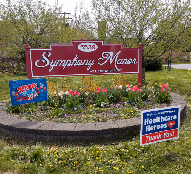 A red sign with white text reading 'Symphony Manor at Lancaster' with the number 5539 above it, surrounded by a circular flower bed with tulips and other flowers. Two smaller signs are placed in the flower bed, one saying 'HEROES WORK HERE' and the other saying 'To Our Essential Workers & Healthcare Heroes Thank You!'. Trees and a grassy area are visible in the background.