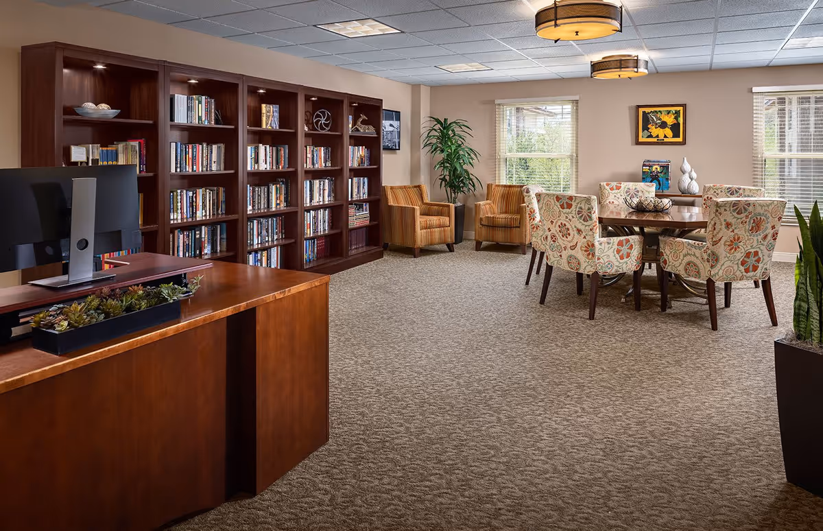 A cozy senior living common area featuring a wooden reception desk with a computer and a planter of succulents, a large bookshelf filled with books, two striped armchairs, a round table surrounded by floral upholstered chairs, potted plants, and windows letting in natural light.