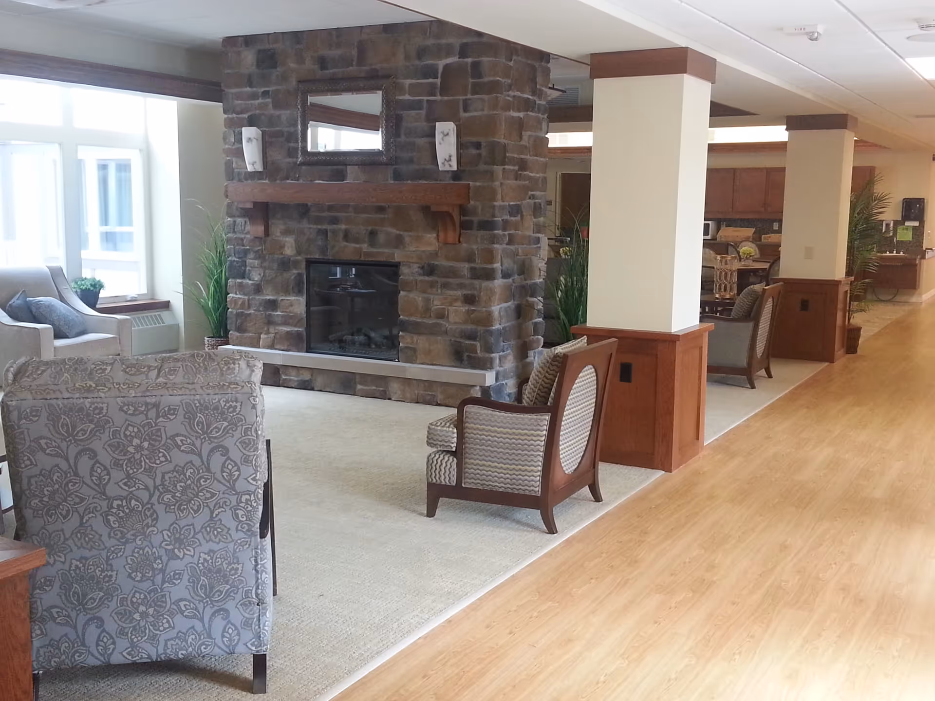A cozy interior common area with a stone fireplace in the center, surrounded by comfortable upholstered chairs and plants. The space has wooden flooring and carpeted seating areas, with large windows letting in natural light.