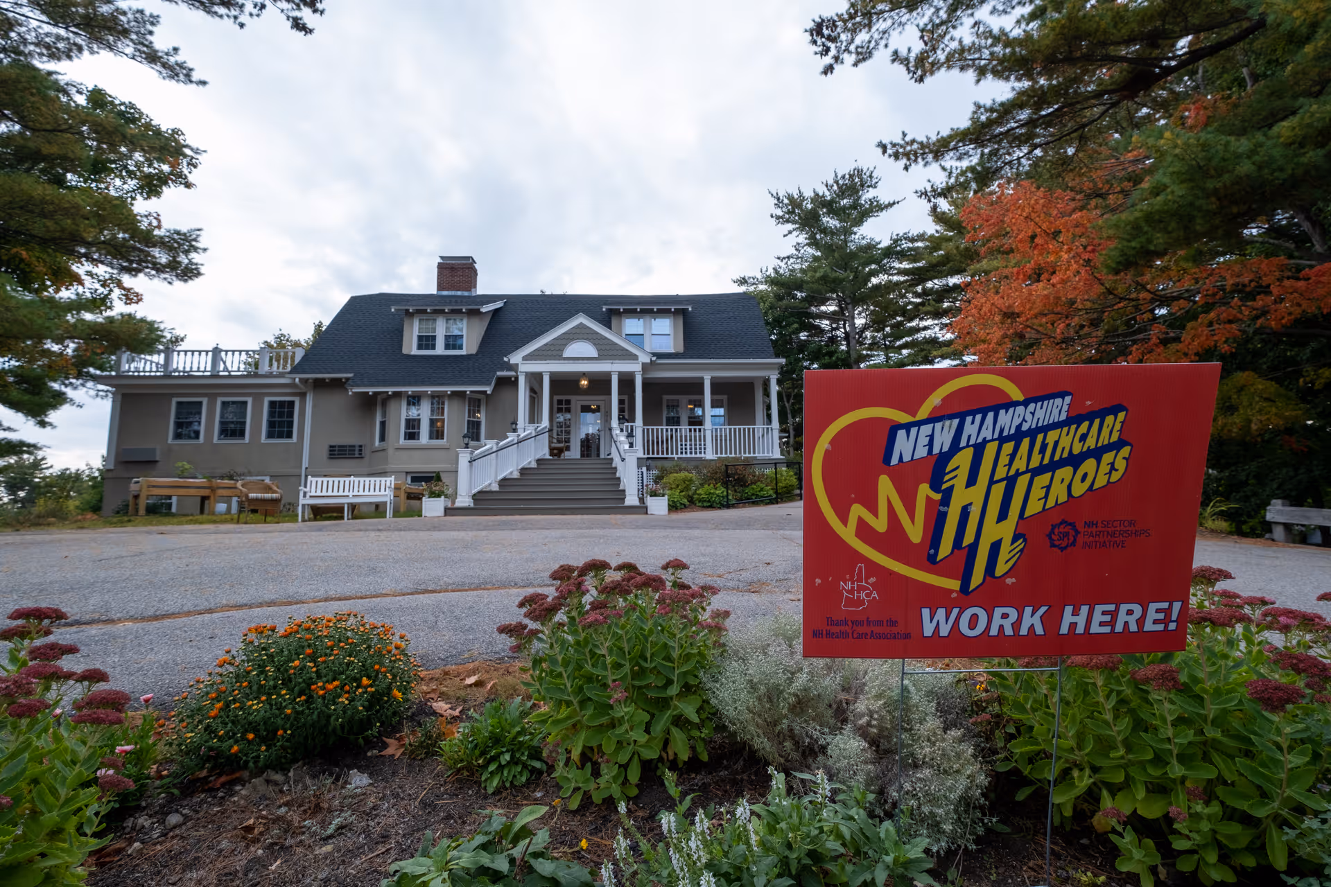 Exterior view of a large house-like building with a front porch and stairs leading up to the entrance. In the foreground, there is a garden with various plants and flowers. A red sign in the garden reads 'New Hampshire Healthcare Heroes WORK HERE!' with a heart and healthcare-themed design. Trees with green and autumn-colored leaves surround the building.