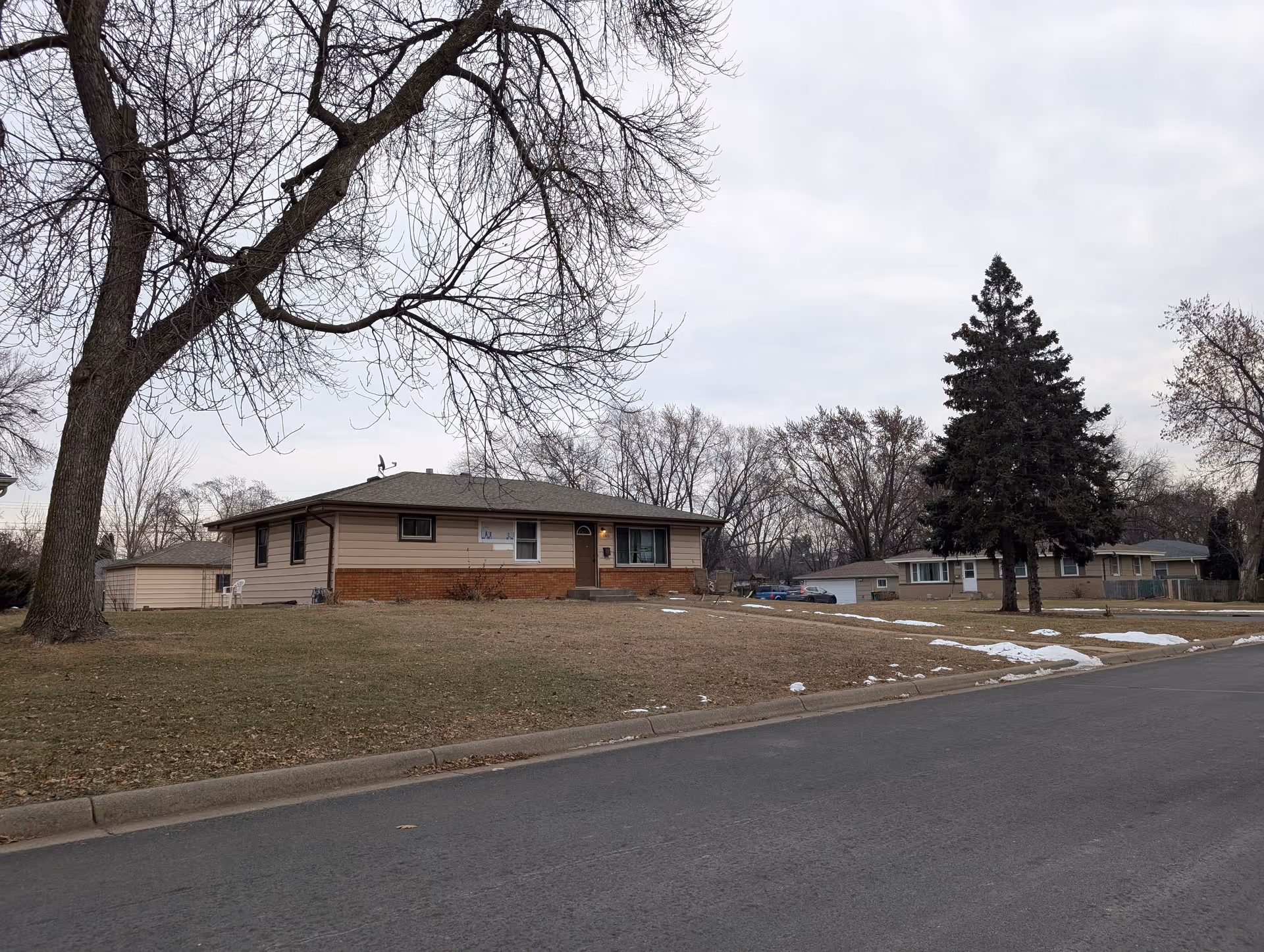 A single-story residential house with beige siding and a brick lower exterior, situated on a slightly elevated grassy lawn with patches of snow. There are leafless trees and a large evergreen tree nearby, with a paved street in the foreground under an overcast sky.