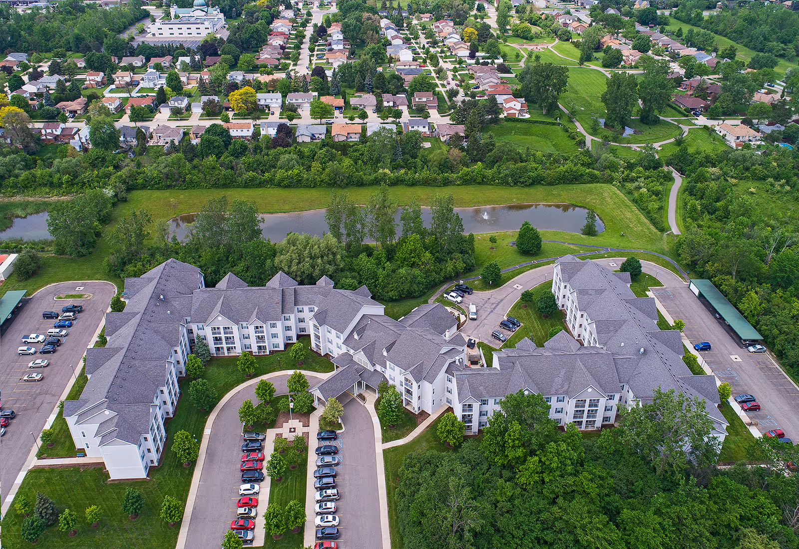 Aerial view of American House Troy senior living facility showing a large multi-wing building surrounded by green lawns, trees, parking lots with cars, a small pond with a fountain, and a residential neighborhood in the background.