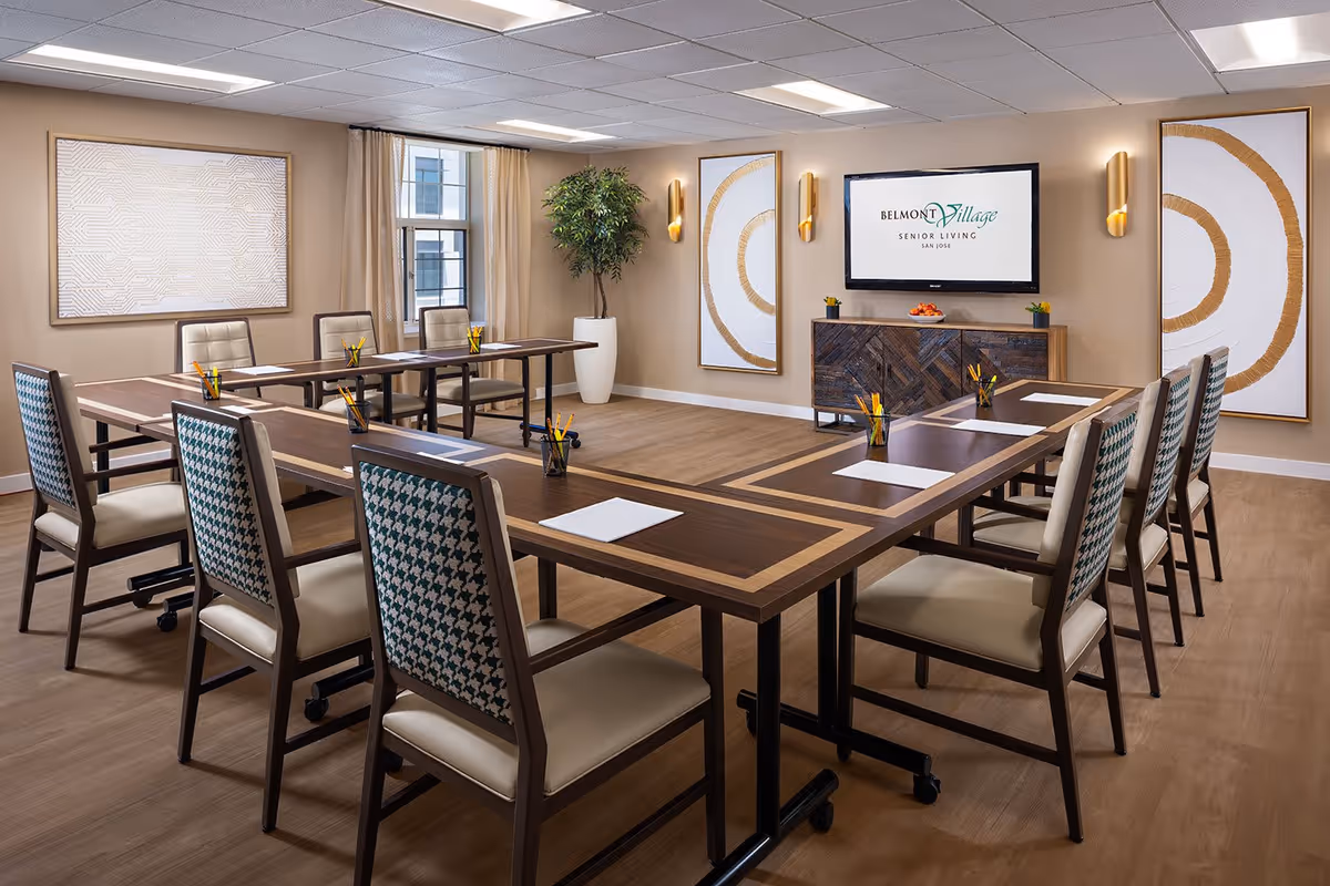 A modern conference or meeting room with a U-shaped arrangement of wooden tables and cushioned chairs. Each table has a notepad and a cup filled with pencils. The room features light-colored walls, three large abstract art pieces with gold accents, a large window with beige curtains, a potted plant, and a flat-screen TV mounted on the wall displaying the Belmont Village Senior Living San Jose logo.