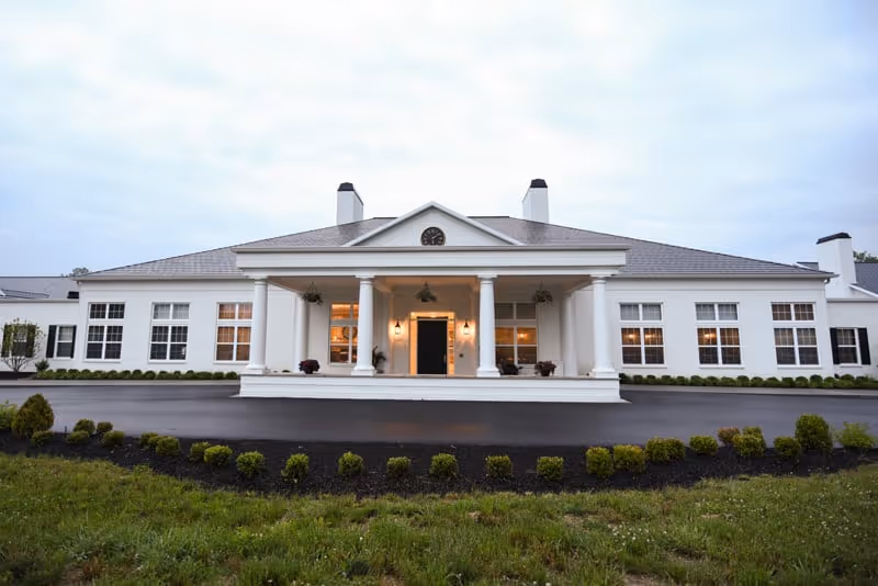 Front exterior view of a large white building with a covered entrance supported by four columns, multiple windows, and a paved driveway surrounded by landscaped bushes and grass under a cloudy sky.