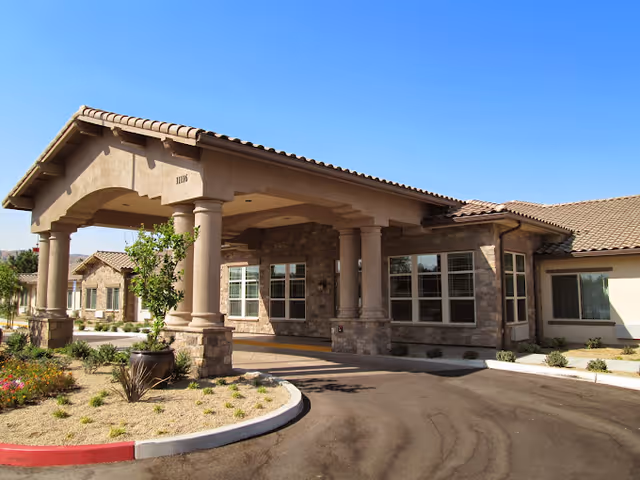 Exterior view of Blossom Grove Alzheimer's Special Care Center showing the entrance with a covered driveway supported by columns, stone and stucco walls, multiple windows, and a landscaped area with desert plants under a clear blue sky.