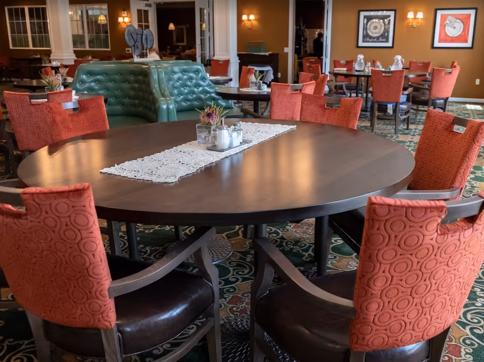 Interior view of a dining room in Ashton Commons featuring a large round wooden table with a white lace runner and small centerpiece. Surrounding the table are red upholstered chairs with circular patterns and dark wooden arms and legs. In the background, there are more tables and chairs, a green tufted booth, wall sconces, framed artwork, and a patterned carpet.