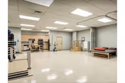 A spacious rehabilitation room with exercise equipment including stationary bikes and weights. The room has a tiled floor, fluorescent ceiling lights, and a treatment table with a red cushion. There are cabinets and a counter area along one wall, and a door leading to another room.