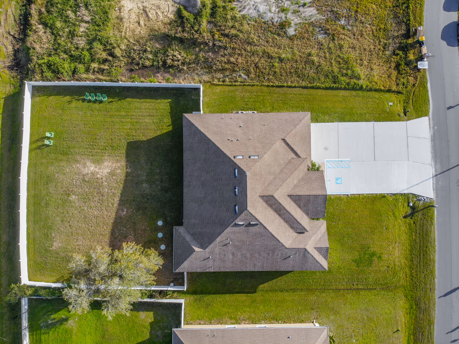 Aerial view of a single building with a brown roof surrounded by green grass and a white fence enclosing a backyard with several green chairs. There is a driveway with a handicapped parking space adjacent to the building.
