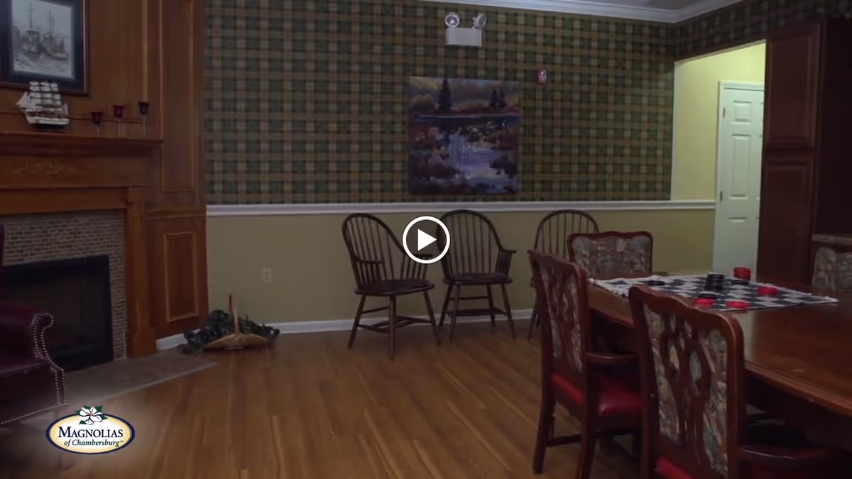 A cozy interior room with wooden flooring, a fireplace with a model ship on the mantle, two wooden chairs against a plaid wallpapered wall, a painting above the chairs, and a wooden dining table with floral upholstered chairs and a checkers game set on top.