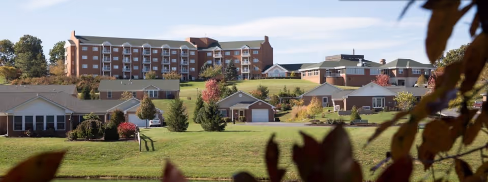 View of a senior living facility named Sunnyside featuring a large multi-story brick building in the background and several single-story houses with garages in the foreground, surrounded by green lawns and trees with autumn foliage.