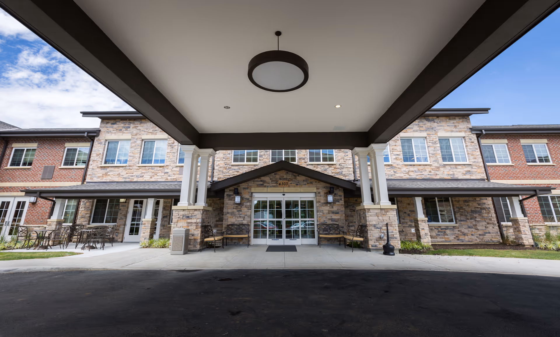 Front entrance of a senior living facility with a covered drop-off area supported by white columns. The building exterior features a combination of brick and stone with multiple windows. There are benches and outdoor seating areas on either side of the entrance under a clear sky.