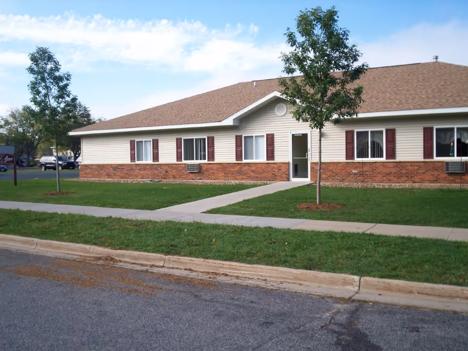 Single-story building with beige siding and red brick lower exterior, several windows with red shutters, a glass door entrance, and a small lawn with two young trees in front under a partly cloudy sky.