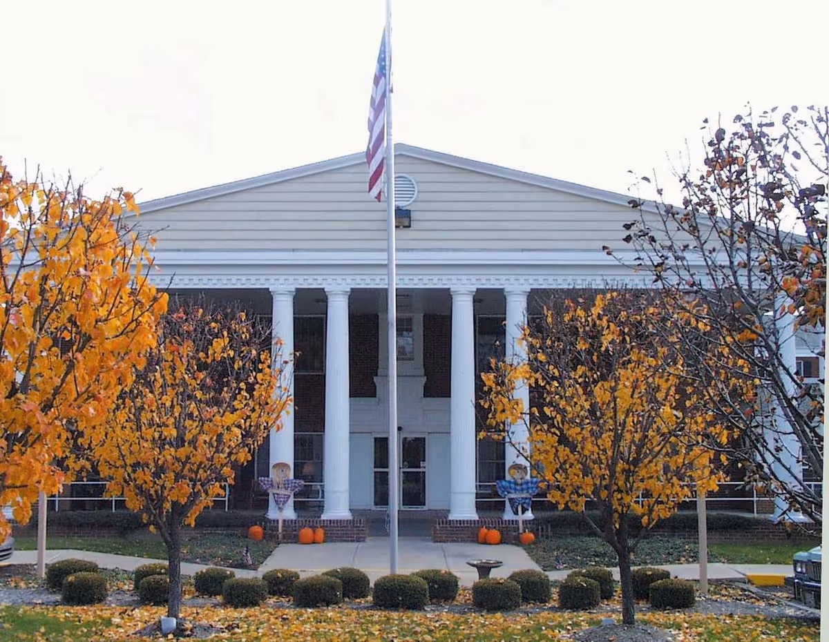 Front exterior view of a building with white columns and a triangular pediment, flanked by trees with orange autumn leaves. An American flag is raised on a flagpole in front of the entrance, and there are pumpkins and scarecrow decorations near the doorway.