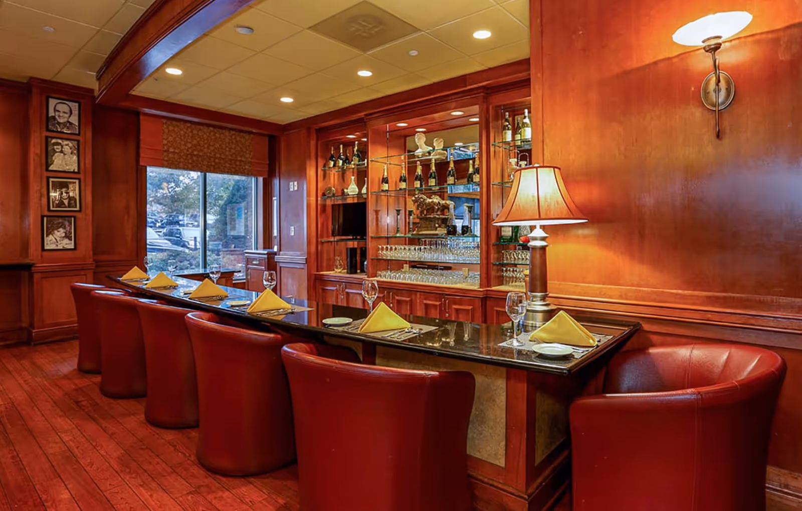 A cozy dining area with a long dark wooden table set with yellow folded napkins, plates, and wine glasses. The table is surrounded by red leather chairs. The room features warm wooden paneling, a glass cabinet displaying bottles and glassware, a table lamp, and a wall sconce. A window with a patterned shade shows an outdoor view.
