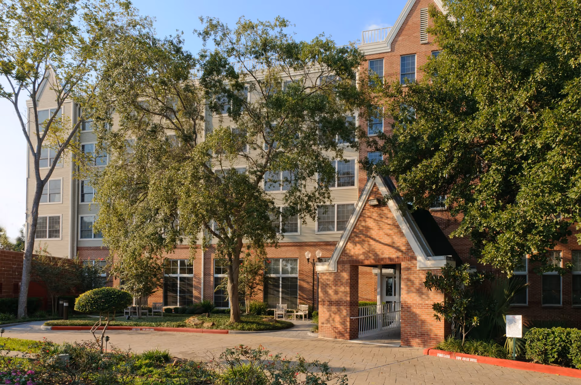 Exterior view of The Vosswood Nursing Center showing a multi-story building with a brick and beige facade, surrounded by trees and greenery. There is a covered entrance with a triangular roof and a paved driveway in front.