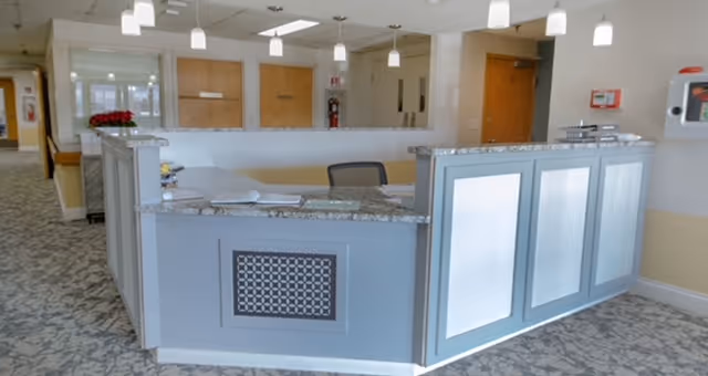 A reception desk area inside a senior living facility with a granite countertop, office chair, and a patterned carpet floor. The background shows wooden doors, a fire extinguisher, and a hallway leading to other rooms.