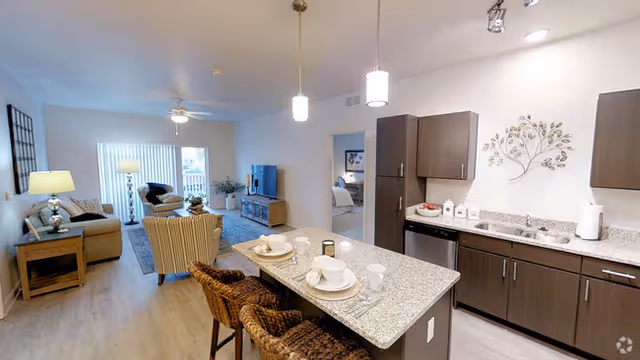 A modern open-concept living space featuring a kitchen with dark wood cabinets, granite countertops, and a breakfast bar with two wicker stools set with plates and glasses. The living room area includes a sofa, armchair, side table with a lamp, and a TV on a stand. Large windows with vertical blinds allow natural light to fill the room. A doorway leads to a bedroom in the background.