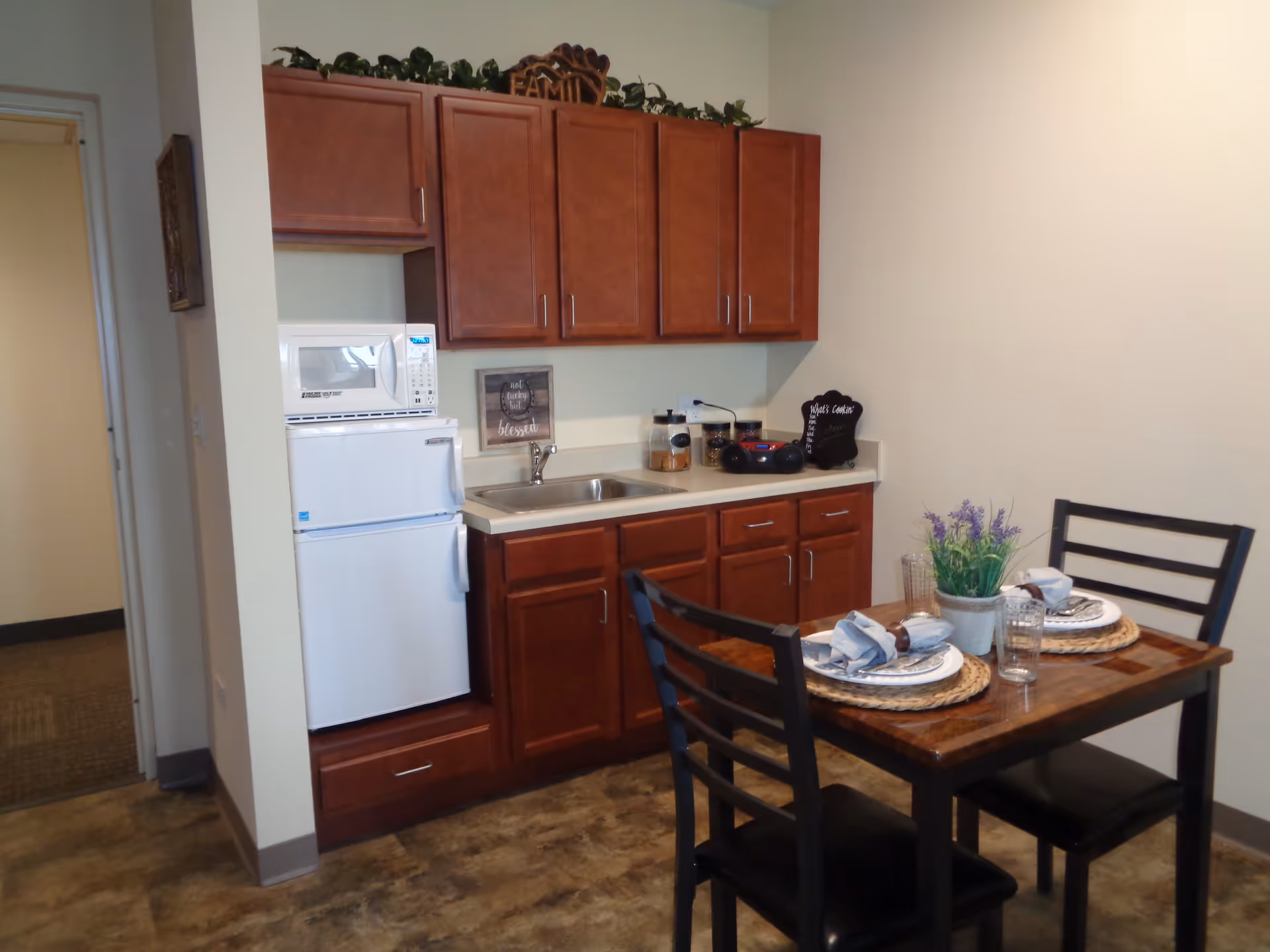 A small kitchen area with wooden cabinets, a white microwave on top of a white mini refrigerator, a sink, and a countertop with jars and a small radio. In front of the kitchen is a wooden dining table set for two with plates, napkins, glasses, and a small potted plant with purple flowers. The floor has a brown tile pattern and the walls are light-colored.