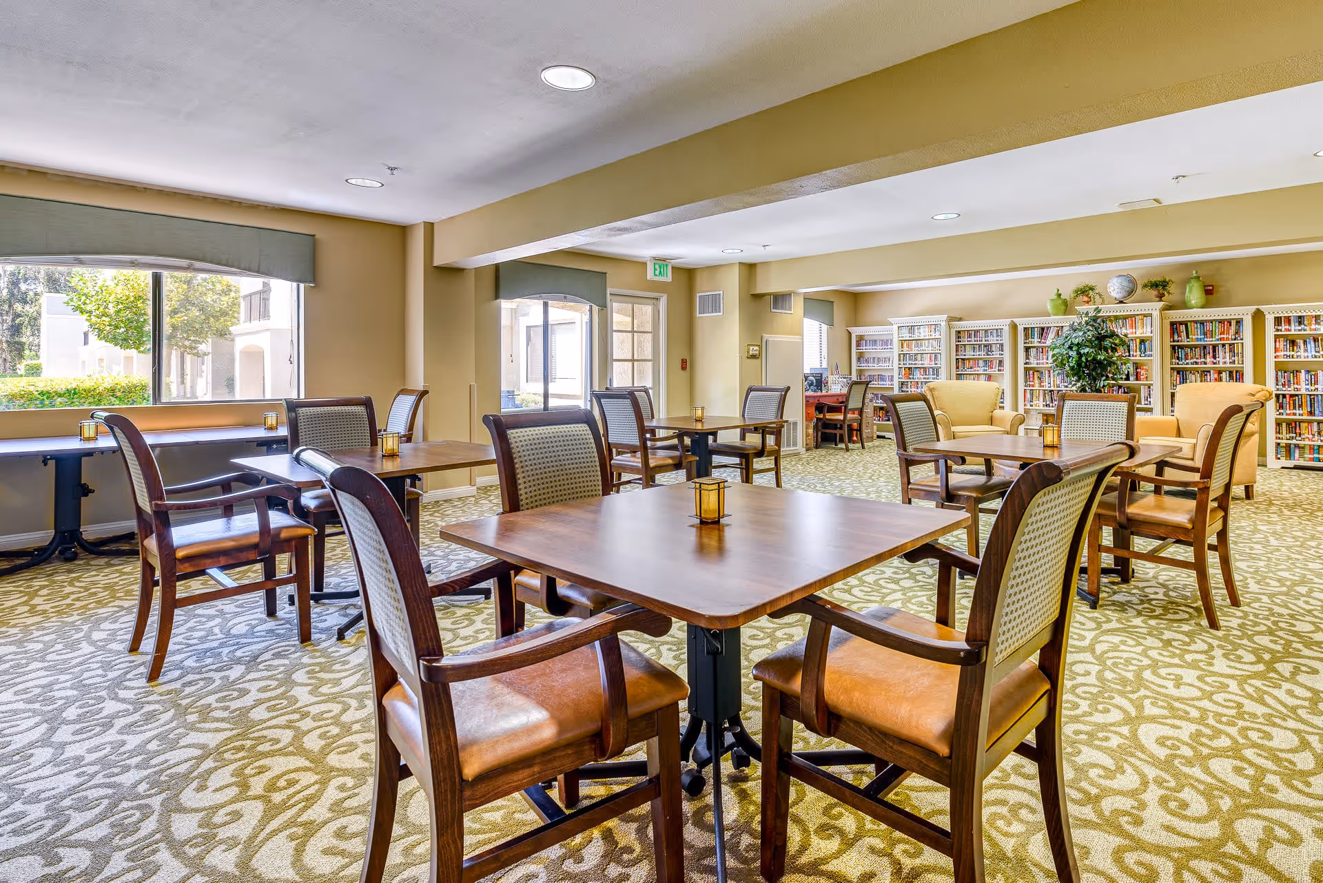 A spacious senior living common area with multiple wooden tables and chairs arranged on a patterned carpet. The room features large windows letting in natural light, bookshelves filled with books along the back wall, and comfortable armchairs near the shelves. The walls are painted a warm beige color, and there are small decorative candles on each table.