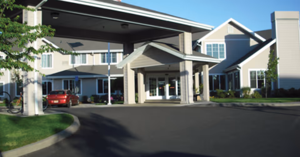 Front exterior view of a senior living facility with a covered entrance, beige siding, white trim, multiple windows, and a paved driveway with a red car parked on the left side.