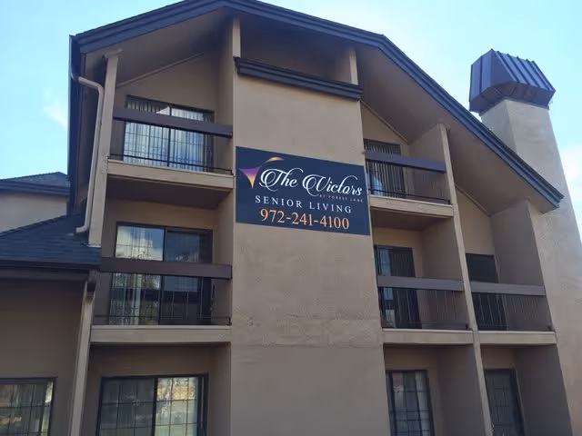 Exterior view of a multi-story senior living building with balconies and a large sign that reads 'The Victors Senior Living 972-241-4100'. The building has beige walls and dark brown railings and roof accents.