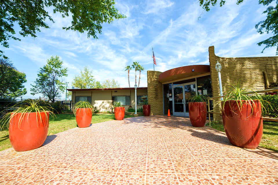 Entrance to Casa Bonita Convalescent Hospital featuring a tiled walkway lined with large red planters containing green plants. The building has a curved brick facade with glass double doors and an American flag flying above. Trees and a clear blue sky are visible in the background.