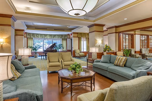 A spacious and well-lit living room area in a senior living facility featuring multiple comfortable sofas and armchairs arranged around a wooden coffee table with a plant centerpiece. The room has wooden flooring, large windows with green curtains, and a grand piano in the background. Warm lighting fixtures and decorative plants add to the cozy atmosphere.